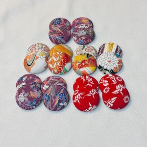 May include: A collection of ten fabric-covered buttons in various colors and patterns. The buttons are arranged in pairs, showcasing floral, geometric, and abstract designs. Colors include red, purple, orange, yellow, and white.