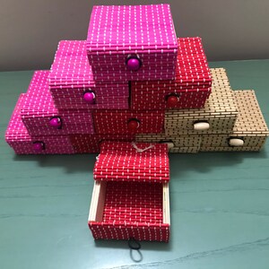 Puede incluir: Una pila de peque&ntilde;as cajas decorativas hechas de bamb&uacute; tejido. Las cajas son en tonos de rosa, rojo y beige. Cada caja tiene un peque&ntilde;o pomo de madera en la parte delantera.