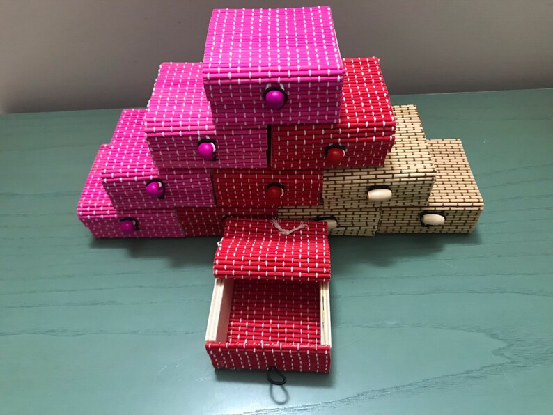 Puede incluir: Una pila de peque&ntilde;as cajas decorativas hechas de bamb&uacute; tejido. Las cajas son en tonos de rosa, rojo y beige. Cada caja tiene un peque&ntilde;o pomo de madera en la parte delantera.