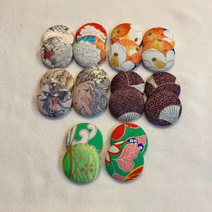 May include: A collection of twelve round fabric-covered buttons. The buttons feature various Japanese-inspired patterns in colors like red, orange, green, and purple. Some have floral designs, while others display geometric or abstract motifs. The buttons are arranged on a white surface.