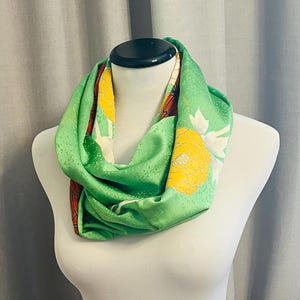 Könnte beinhalten: Ein grüner Seidenschal mit einem floralen Muster in Gelb, Weiß und Rot. Der Schal ist gefaltet und auf einer weißen Schaufensterpuppe ausgestellt.