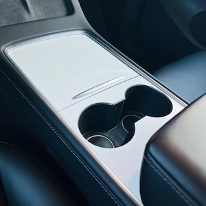 May include: A white center console cover with a smooth finish installed on a vehicle interior. The cover features a rectangular sliding panel section and a dual cup holder cutout, integrated into the black leather-trimmed console area.