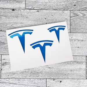 May include: Three blue Tesla logo decals on a white background. The decals are in the shape of a stylized "T" with a horizontal bar at the top. The decals are placed on a light gray wood-grain surface.