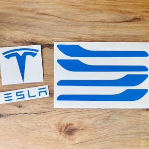 May include: Blue Tesla logo sticker, Tesla text sticker, and four blue door handle stickers on a white background. The stickers are on a wooden surface.