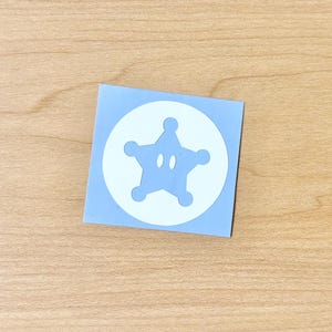 以下が含まれることがあります： 中央に白い円が配置された正方形のライトブルーのステッカー。白い円の中には、角が丸みを帯びたライトブルーの五芒星があり、星の中央には目のような形をした2つの小さな楕円形の切り抜きが施されています。