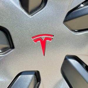 May include: Close-up of a silver car wheel cover with a red Tesla logo in the center. The logo is a stylized letter "T". The wheel cover has a star-like design with black accents.