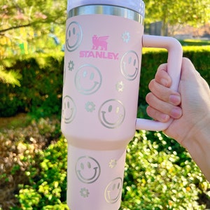 May include: A pink stainless steel travel mug with a handle. The mug has a white lid and is decorated with silver smiley faces and flowers. The mug has the word "STANLEY" printed on it.