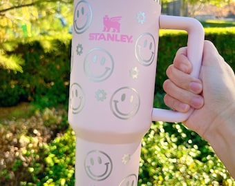 May include: A light pink Stanley tumbler with a handle and a lid, featuring a repeating pattern of silver-colored smiley faces and small flower icons. The Stanley logo is printed in pink on the upper portion of the cup, centered above the pattern.