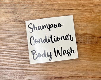 May include: A square white adhesive decal featuring the black text Shampoo, Conditioner, and Body Wash in a cursive script font. The lettering is arranged in three distinct lines, providing clear labels for bathroom organization and storage.