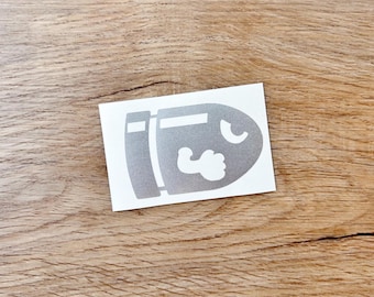 May include: A rectangular white sticker featuring a metallic silver graphic of a bullet character with a flexed arm and a single eye. The design is flat and centered on the white rectangular backing, showcasing a stylized cartoon aesthetic.