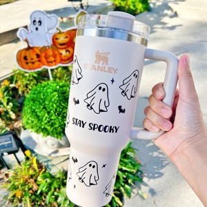 May include: A light pink Stanley tumbler with a handle and a clear lid, featuring black graphic prints of ghosts, small bats, and stars. The text "STAY SPOOKY" is printed in black on the front of the tumbler, positioned below the ghost illustrations.