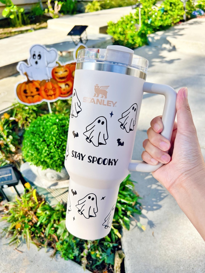 May include: A light pink Stanley tumbler with a handle and a clear lid, featuring black graphic prints of ghosts, small bats, and stars. The text "STAY SPOOKY" is printed in black on the front of the tumbler, positioned below the ghost illustrations.