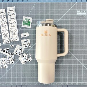 May include: A cream-colored Stanley tumbler with a handle and lid, accompanied by a collection of small ghost-themed vinyl decals, a Curad alcohol prep pad, and a sticker featuring the text STAY SPOOKY, all arranged on a gray cutting mat.