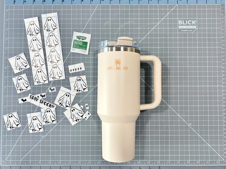 May include: A cream-colored Stanley tumbler with a handle and lid, accompanied by a collection of small ghost-themed vinyl decals, a Curad alcohol prep pad, and a sticker featuring the text STAY SPOOKY, all arranged on a gray cutting mat.