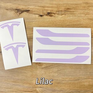以下が含まれることがあります： 白い台紙に貼られたライラック色のビニール製デカールセット。テスラのロゴエンブレム2つと、ハンドル状の細長いストリップ4つが含まれています。画像中央下部には白い文字で「Lilac」と印字されており、デカールの色を示しています。