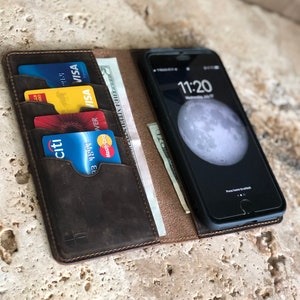 K&ouml;nnte beinhalten: Eine braune Ledergeldb&ouml;rse mit einem integrierten Handy-Etui. Die Geldb&ouml;rse hat mehrere Kartenf&auml;cher und ein Fach f&uuml;r Bargeld. Das Handy-Etui hat einen schwarzen Bildschirm mit einem wei&szlig;en Mond und die Uhrzeit 11:20 angezeigt.