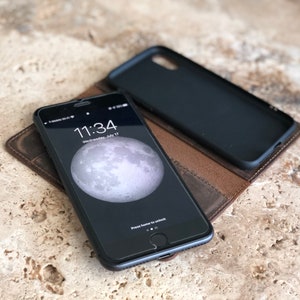 K&ouml;nnte beinhalten: Eine braune Leder-Handyh&uuml;lle mit einem schwarzen Telefon darin. Der Telefonbildschirm zeigt die Uhrzeit 11:34 und das Datum Mittwoch, 17. Juli. Die Handyh&uuml;lle hat eine Klappe, die &uuml;ber das Telefon geklappt wird.