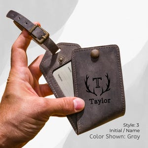 May include: Gray leather luggage tag with a buckle strap. The tag features a card holder and a cover with the initial "T" and the name "Taylor" printed below. The text "Style: 3, Initial / Name, Color Shown: Gray" is also visible.