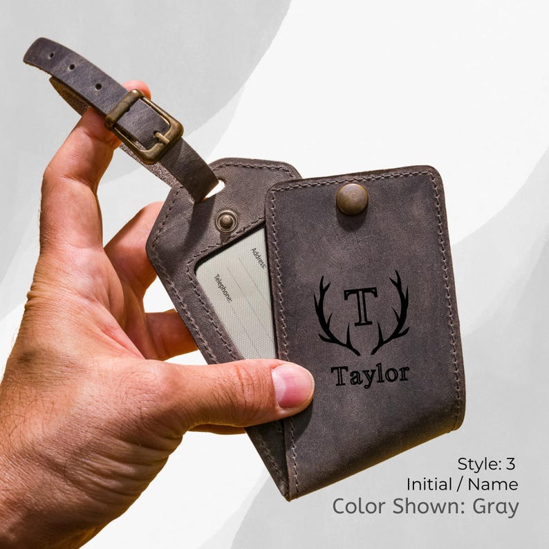 May include: Gray leather luggage tag with a buckle strap. The tag features a card holder and a cover with the initial "T" and the name "Taylor" printed below. The text "Style: 3, Initial / Name, Color Shown: Gray" is also visible.