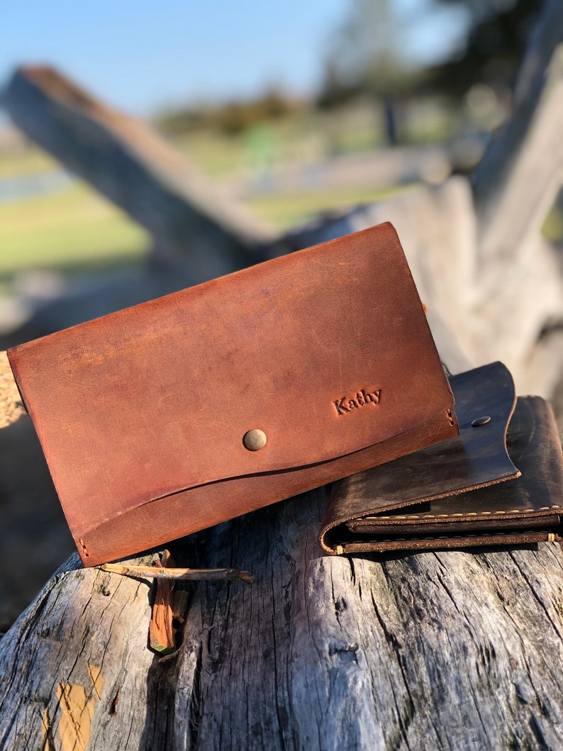 May include: Two brown leather wallets, one with the name "Kathy" embossed on the front. The wallets are sitting on a weathered wooden surface.