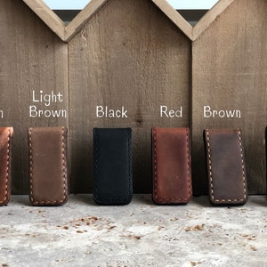 May include: Seven leather money clips in different colors: tan, light brown, black, red, brown, and gray. The clips are arranged in a row on a wooden surface.