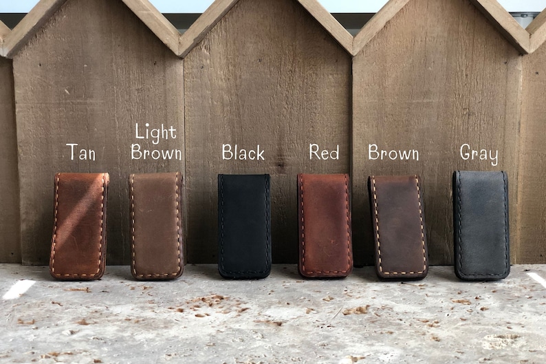 May include: Seven leather money clips in different colors: tan, light brown, black, red, brown, and gray. The clips are arranged in a row on a wooden surface.