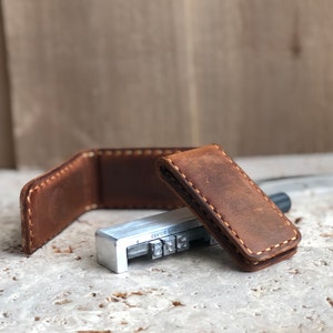 May include: A brown leather money clip with white stitching. The clip is closed and sitting on a light gray surface. A metal tool with the word "CENTER" is visible in the background.