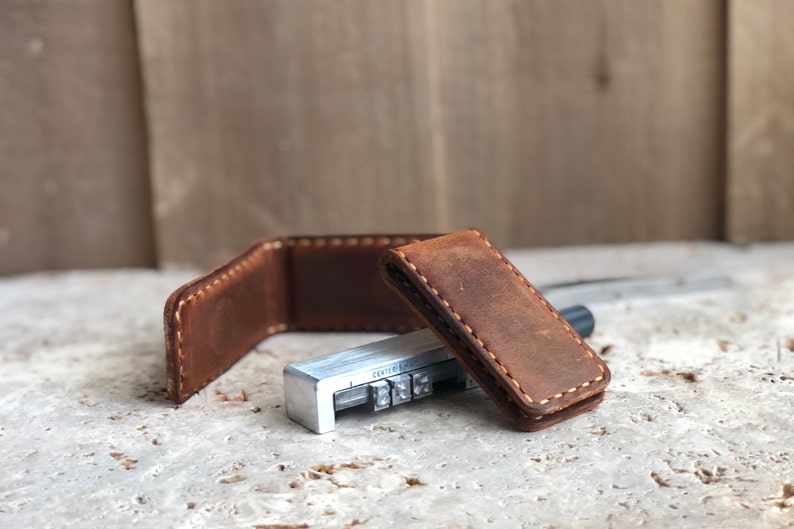 May include: A brown leather money clip with white stitching. The clip is closed and sitting on a light gray surface. A metal tool with the word "CENTER" is visible in the background.