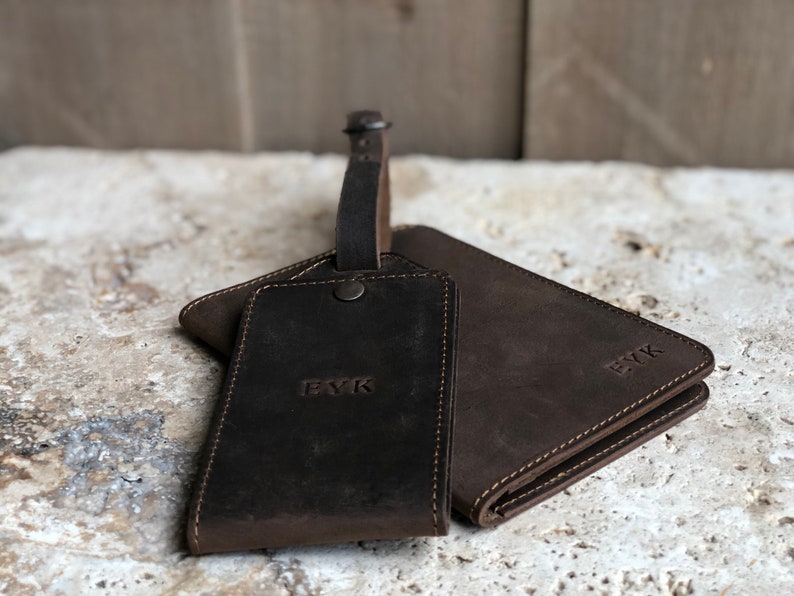 Puede incluir: Un conjunto de accesorios de viaje de cuero marr&oacute;n oscuro, que incluye una etiqueta de equipaje con correa y una cartera. Ambos art&iacute;culos est&aacute;n grabados con las iniciales "EYK". El cuero tiene un aspecto texturizado y los art&iacute;culos est&aacute;n cosidos con un hilo m&aacute;s claro.