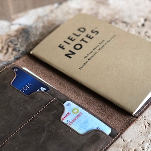 May include: A brown leather wallet with a credit card and a driver's license visible in the card slots. A Field Notes memo book rests on top. The memo book is tan with black text that reads "FIELD NOTES".