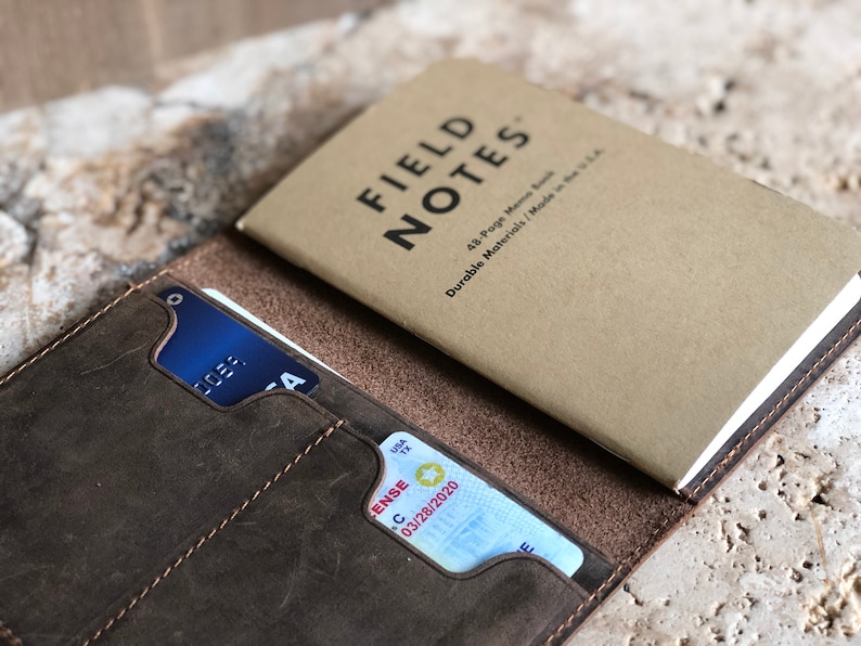 May include: A brown leather wallet with a credit card and a driver's license visible in the card slots. A Field Notes memo book rests on top. The memo book is tan with black text that reads "FIELD NOTES".