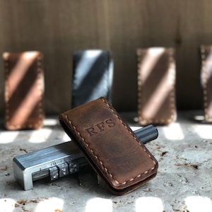 May include: A brown leather money clip with the initials "RFS" embossed on the front. The clip is sitting on a concrete surface next to a metal tool with letter stamps.