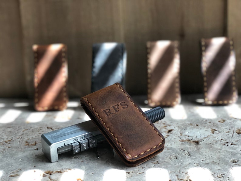 May include: A brown leather money clip with the initials "RFS" embossed on the front. The clip is sitting on a concrete surface next to a metal tool with letter stamps.