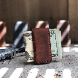 May include: A brown leather money clip with a stitched edge, holding a stack of US dollar bills. The clip is on a gray surface with light and dark stripes.