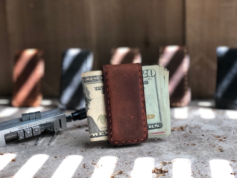 May include: A brown leather money clip with a stitched edge, holding a stack of US dollar bills. The clip is on a gray surface with light and dark stripes.