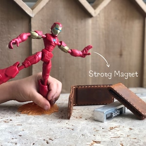 May include: A red Iron Man action figure with a strong magnet in its foot, holding onto a brown leather wallet with a silver metal object inside.