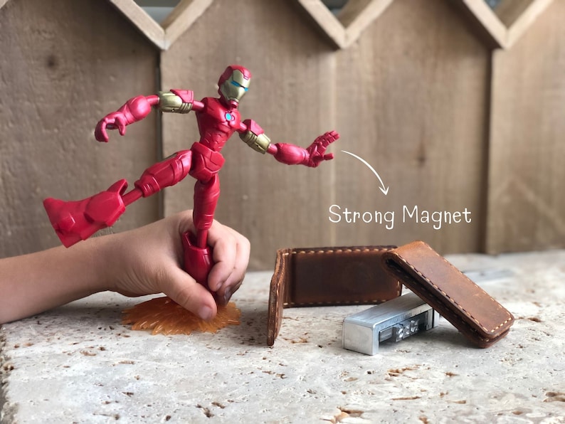 May include: A red Iron Man action figure with a strong magnet in its foot, holding onto a brown leather wallet with a silver metal object inside.