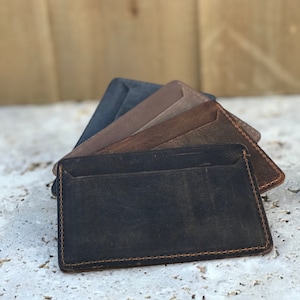 May include: A stack of five leather wallets in different shades of brown and black. The wallets are made from soft, supple leather and have a simple, minimalist design.
