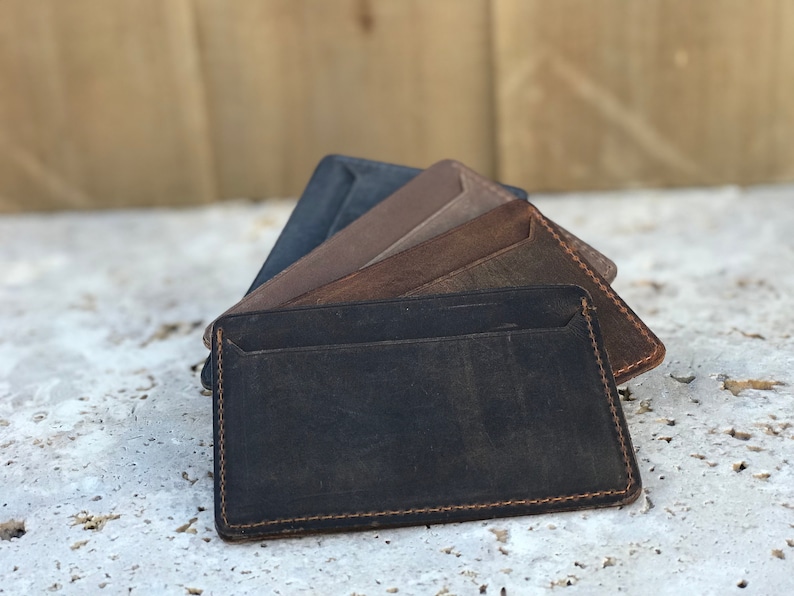 May include: A stack of five leather wallets in different shades of brown and black. The wallets are made from soft, supple leather and have a simple, minimalist design.