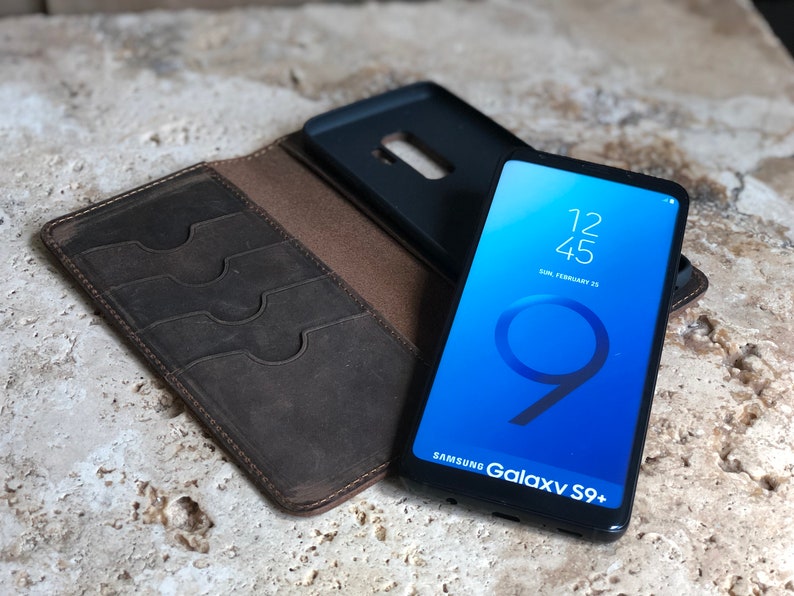 K&ouml;nnte beinhalten: Ein braunes Leder-Telefonetui mit Steckpl&auml;tzen f&uuml;r Karten und ein schwarzes Telefonetui f&uuml;r ein Samsung Galaxy S9+ Telefon. Der Telefonbildschirm zeigt die Uhrzeit 12:45 und das Datum Sonntag, 25. Februar.