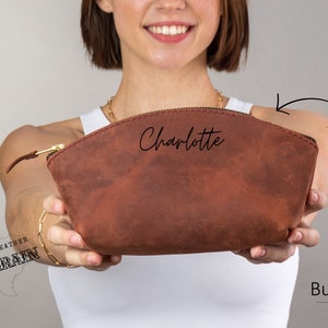 May include: Burgundy leather makeup bag with a zipper closure and the name "Charlotte" written in cursive on the front. The bag is made of full grain leather.