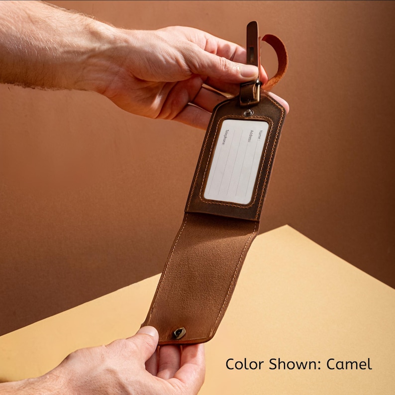 May include: Camel-colored leather luggage tag with a rectangular window for name, address, and phone number. The tag has a buckle closure and a leather strap for attachment. The text "Color Shown: Camel" is visible.