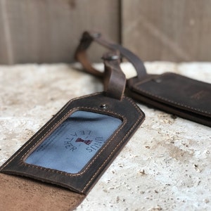 May include: Two brown leather luggage tags with a distressed finish. One tag is open, revealing a clear window with a blue and white striped background and the text "UNUS LEATHER". The other tag is closed. Both have leather straps.
