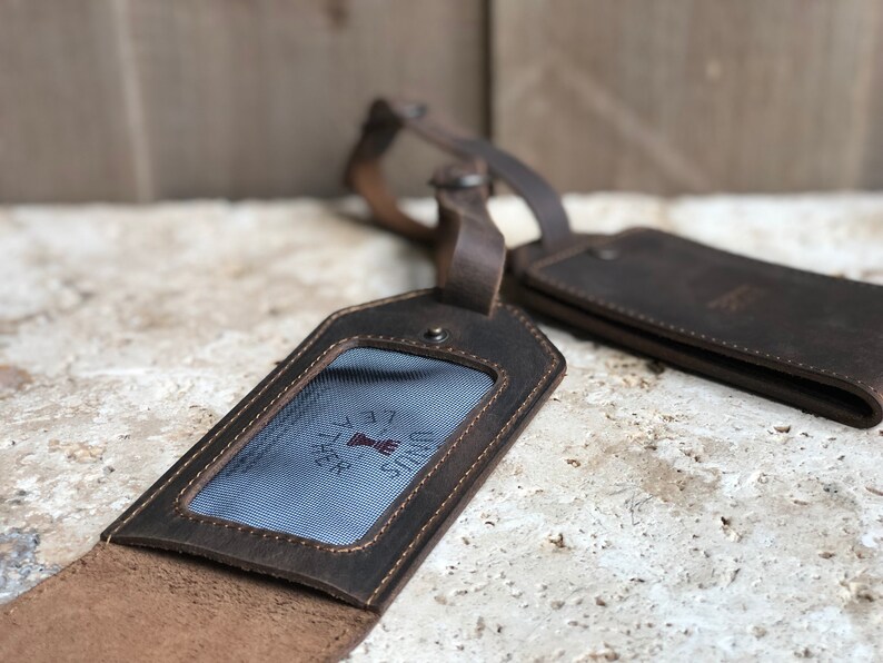 May include: Two brown leather luggage tags with a distressed finish. One tag is open, revealing a clear window with a blue and white striped background and the text "UNUS LEATHER". The other tag is closed. Both have leather straps.