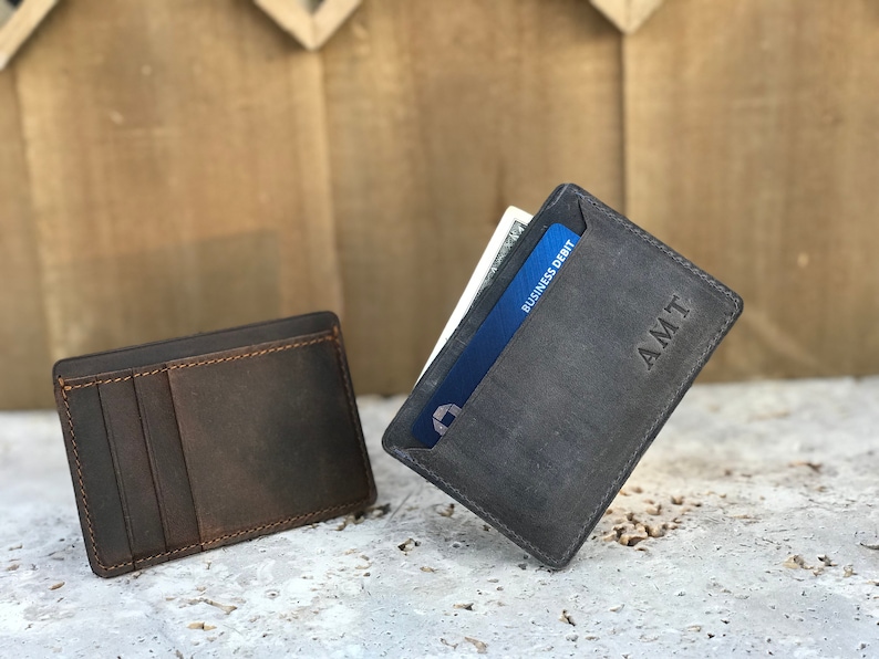 May include: Two brown leather card holders. One is closed and the other is open with a blue credit card and cash inside. The open card holder has the initials "AMT" embossed on the front.