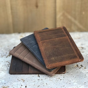 May include: Four leather card holders in different colours: brown, tan, grey, and dark blue. The card holders are stacked on top of each other.