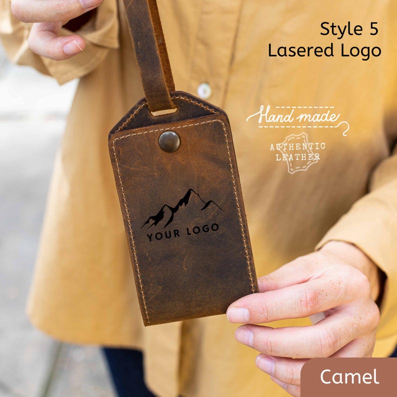 May include: A camel-colored leather luggage tag with a mountain logo and the text "YOUR LOGO." The tag is labeled "Hand made" and "AUTHENTIC LEATHER." The tag has a strap and a button closure. The style is "Lasered Logo."