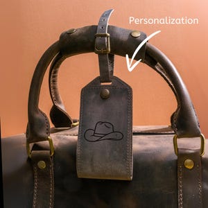 May include: A dark brown leather travel bag with a matching handle and a personalized luggage tag. The tag features a cowboy hat design. The bag has brass-colored hardware and a buckle closure.