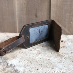 Puede incluir: Una etiqueta de equipaje de cuero marr&oacute;n con una correa de hebilla. La etiqueta tiene una ventana rectangular con el texto "UNUS LEATHER" y un logotipo. La etiqueta est&aacute; abierta, revelando un interior azul claro.