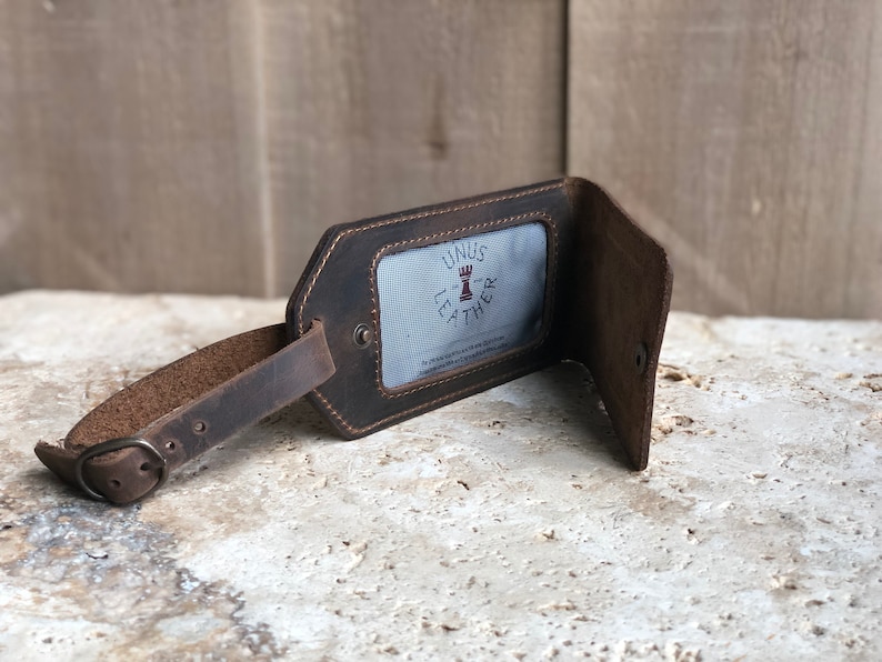 Puede incluir: Una etiqueta de equipaje de cuero marr&oacute;n con una correa de hebilla. La etiqueta tiene una ventana rectangular con el texto "UNUS LEATHER" y un logotipo. La etiqueta est&aacute; abierta, revelando un interior azul claro.